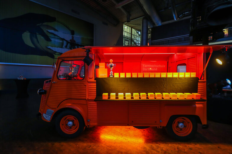 Orange, old transport car is parked openly in an event hall and has various prizes set up in the interior, which are illuminated in orange.