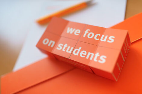 Photo of an opened folding cube with the imprint "we focus on students" lies on documents. __ Opened folding cube with the imprint "we focus on students" lies on documents.