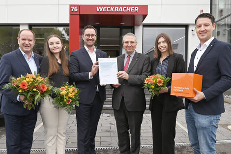Six people stand next to each other, looking friendly into the camera, in the background a short flight of stairs leading to the entrance of a store with the word "Weckbacher" on the canopy. Three people hold a bouquet of flowers in their hands, the two in the middle hold a certificate up to the camera.