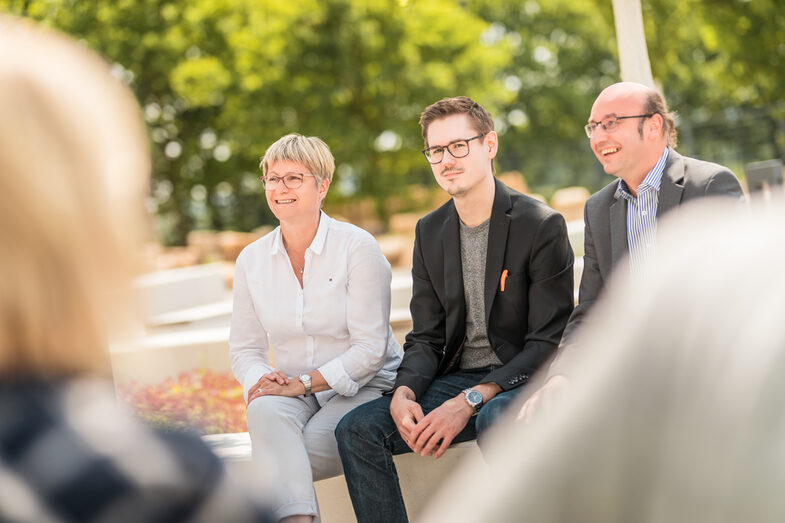Photo of three people sitting outside on campus. A doctoral student in the middle, next to him two colleagues who are supervising him during his doctorate.