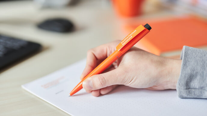 Photo of a hand writing on a notepad with a ballpoint pen. The pen is orange and bears the University of Applied Sciences lettering in white.