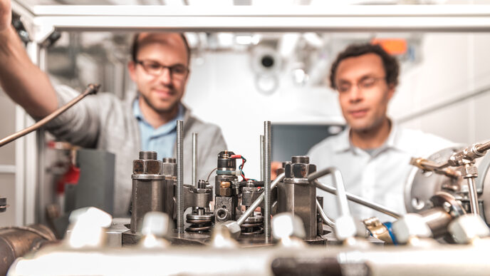 Photo of elements of a motor assembly. Behind it you can see a doctoral student and his professor looking at the engine.