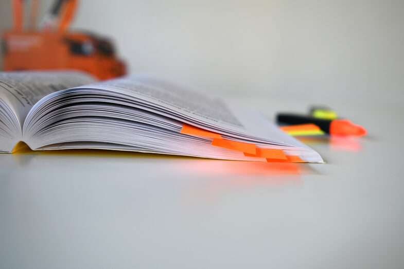 Photo of an open book with orange sticky notes inside. In the background is a pencil box and highlighters.